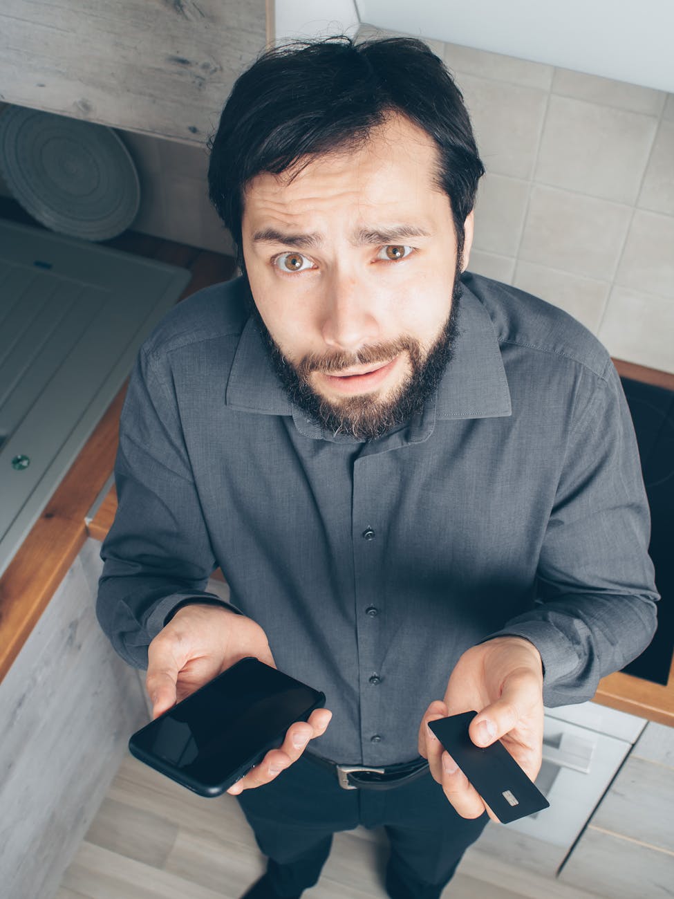 A confused man indoors looks up while holding a smartphone and credit card, concerned about a financial issue.
