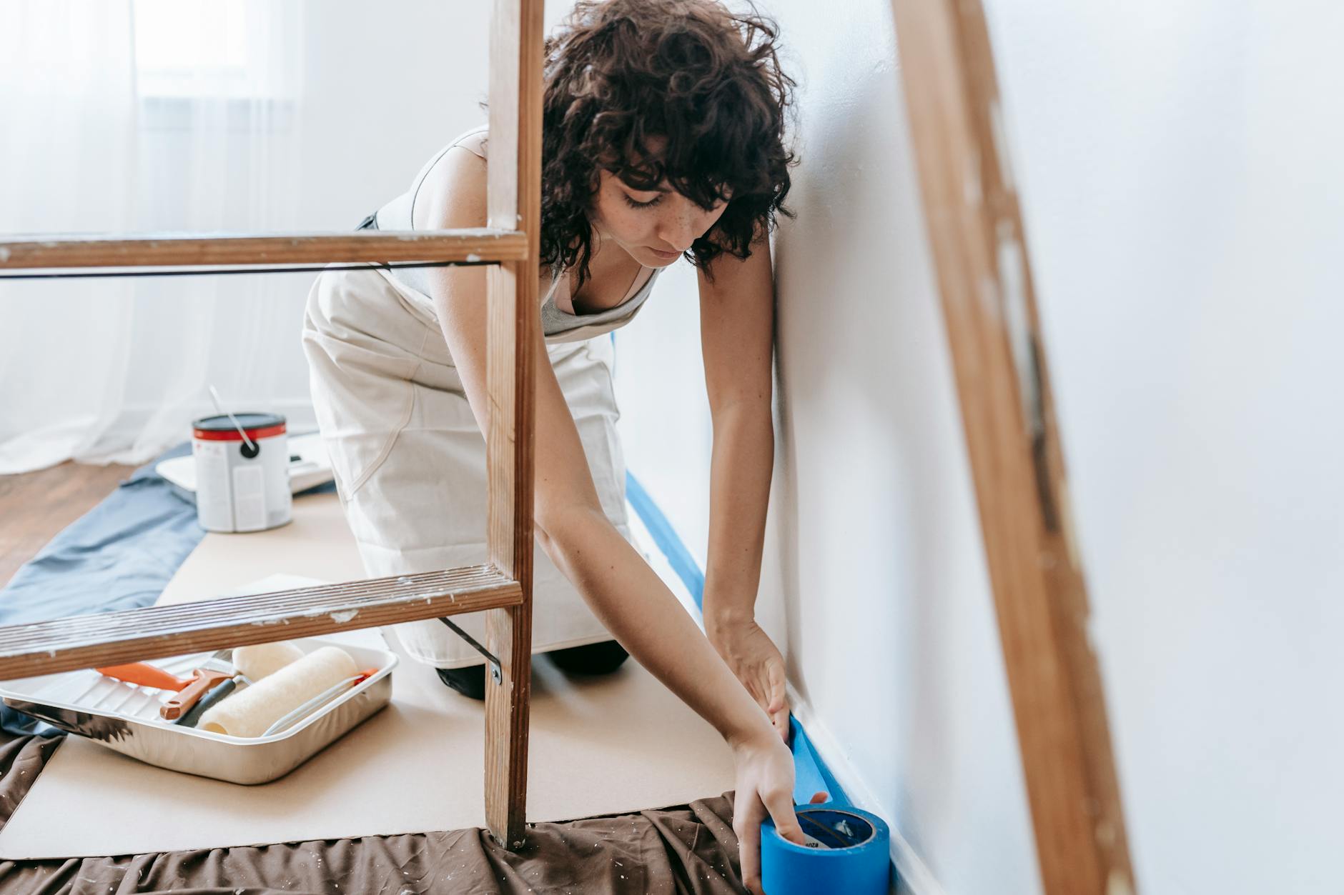 Woman uses tape to prep wall for painting, part of a DIY home renovation project indoors.